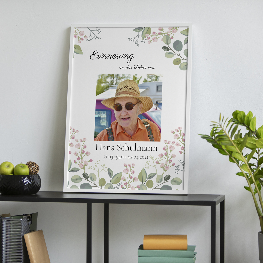 Trauerposter mit Porträt und zarten Blumen im weissen Rahmen auf einer schwarzen Konsole neben einer Pflanze